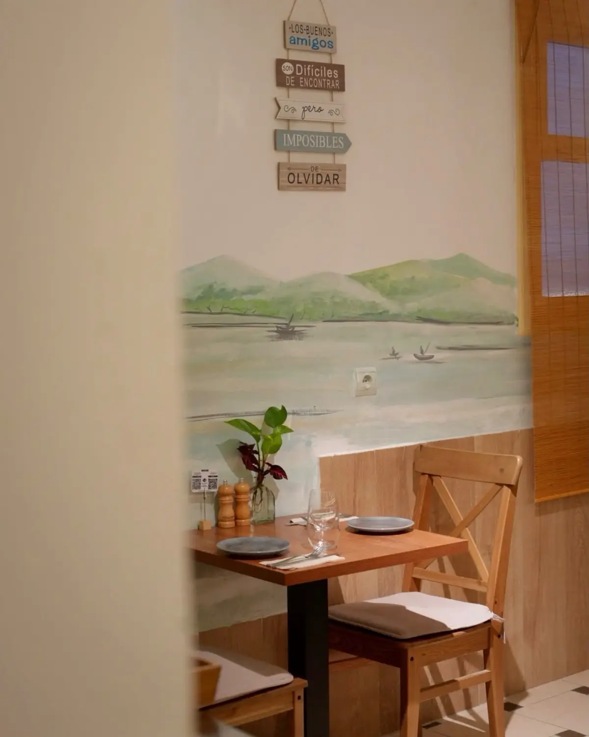 A table for two under a painted lake, with the sign “Los buenos amigos son difíciles de encontrar, pero imposibles de olvidar”