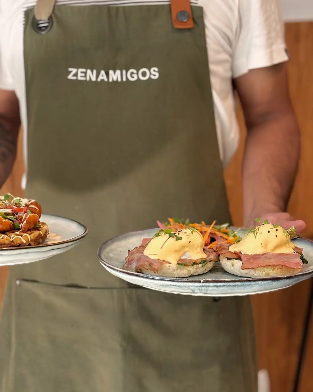 A cook in a ZENAMIGOS apron carrying a Classic Benny and a karaage waffle